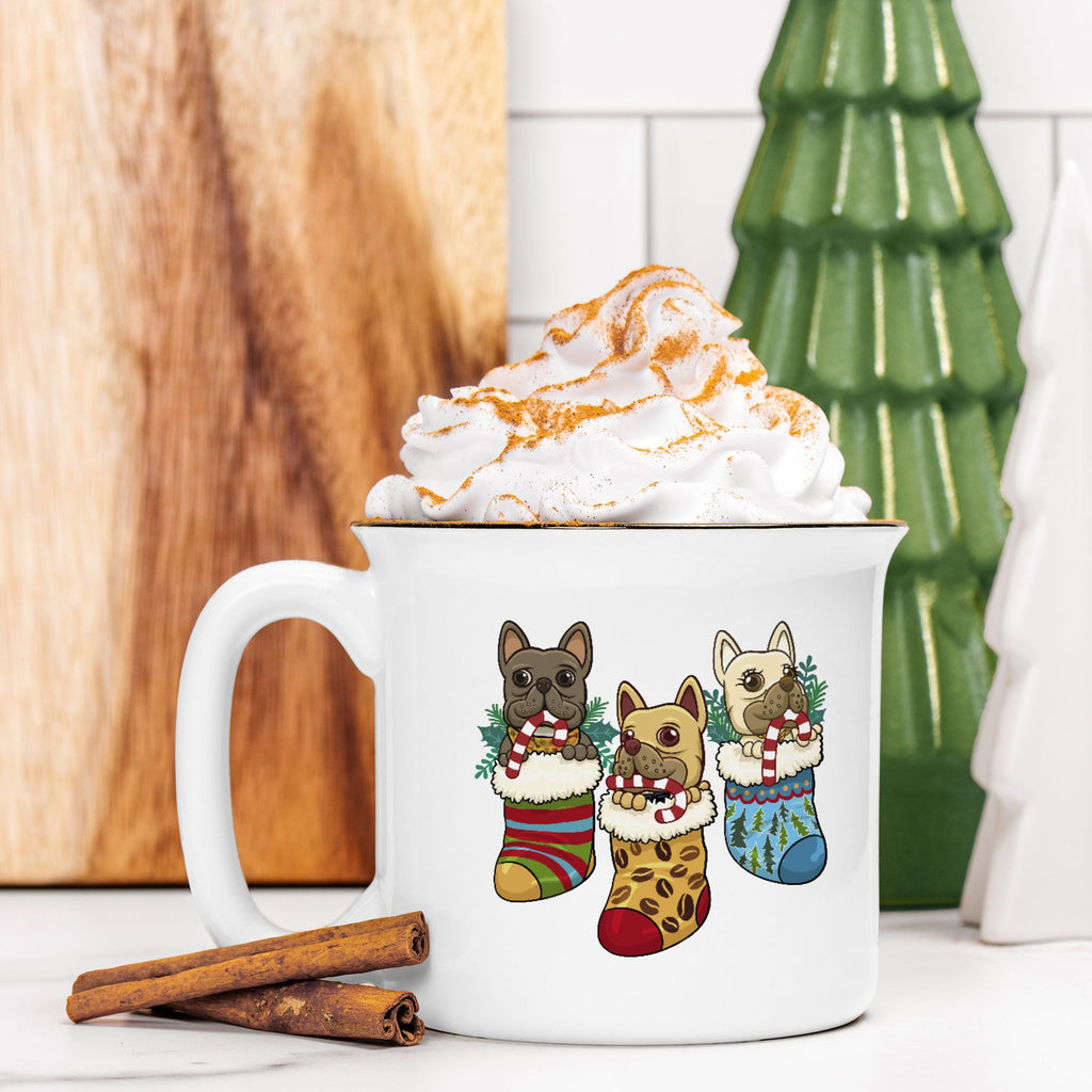White mug with Christmas-themed dog illustrations filled with hot chocolate and whipped cream, surrounded by cinnamon sticks and a green ceramic tree.